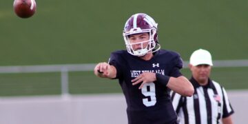 Quarterback in black uniform throws a football during a game, with a referee in stripes in the background.