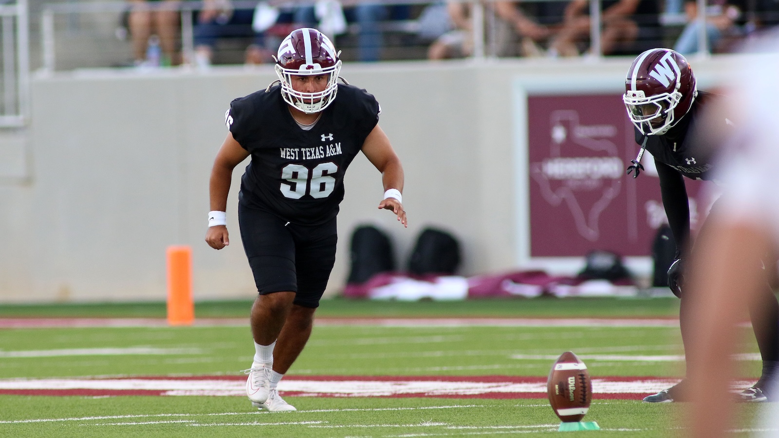 West Texas A&M football player #96 in a black uniform crouches on the field beside the ball on a tee, ready to move; another player in the background.