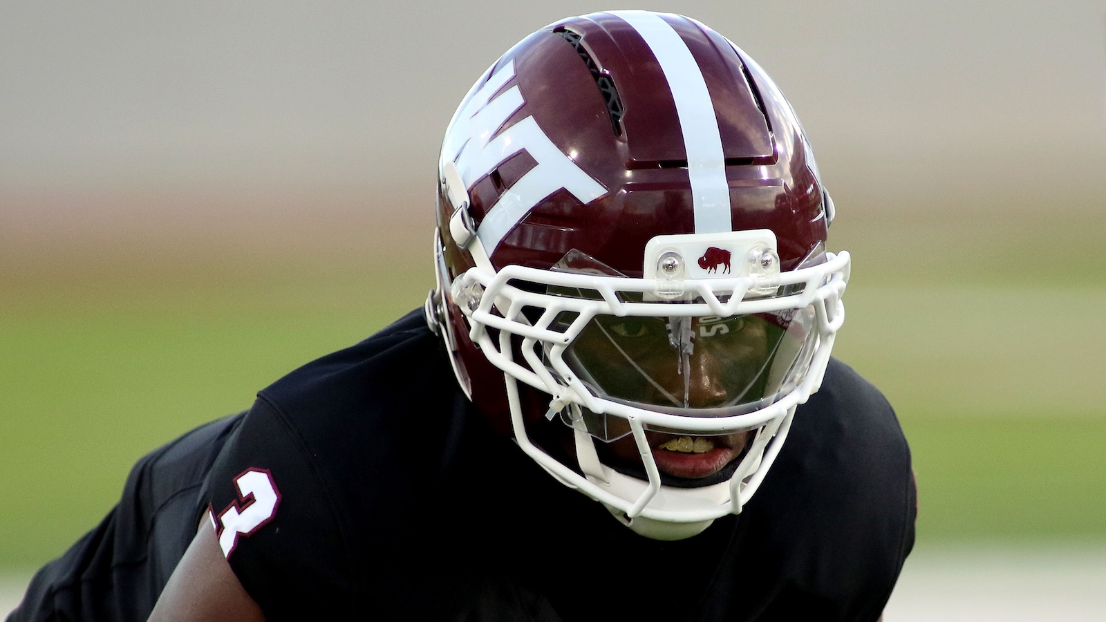 Close-up of a football player wearing a maroon helmet with a white stripe and facemask, focused on the field in a black jersey.