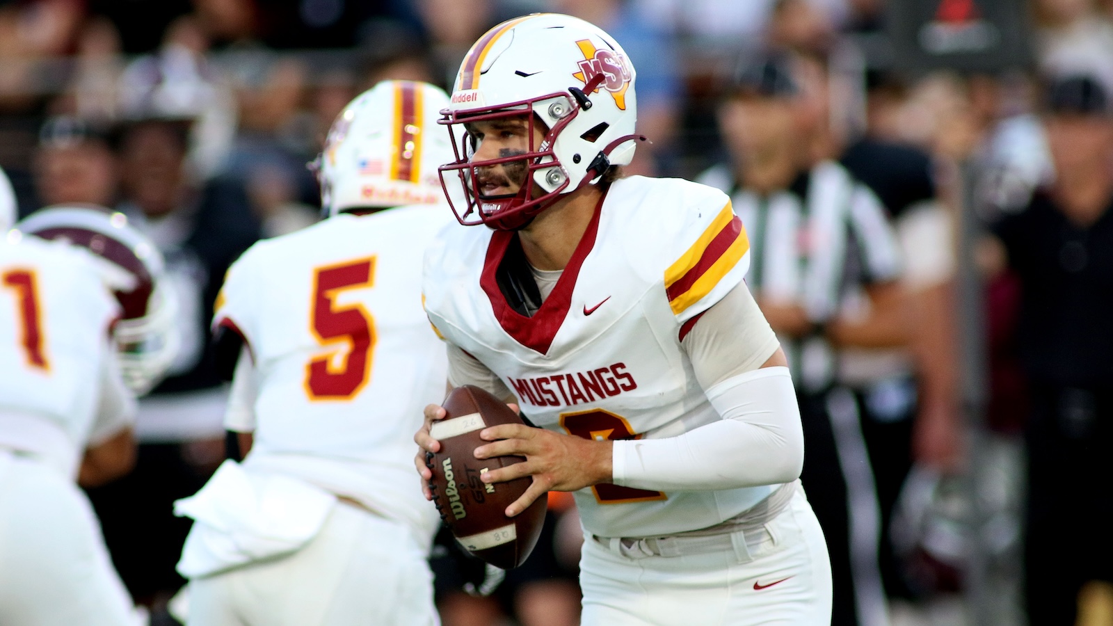 Quarterback in a white Mustangs uniform grips a football during a game, ready to pass on the field.