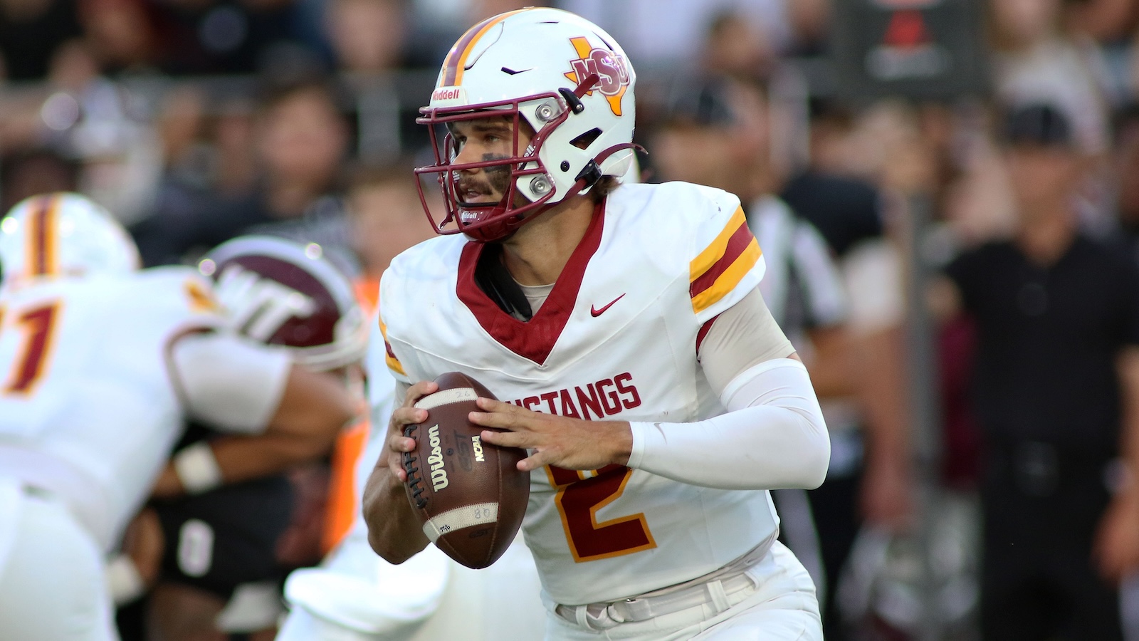 Quarterback in a white Mustangs uniform with number 2 holding a football and scanning the field during a game.