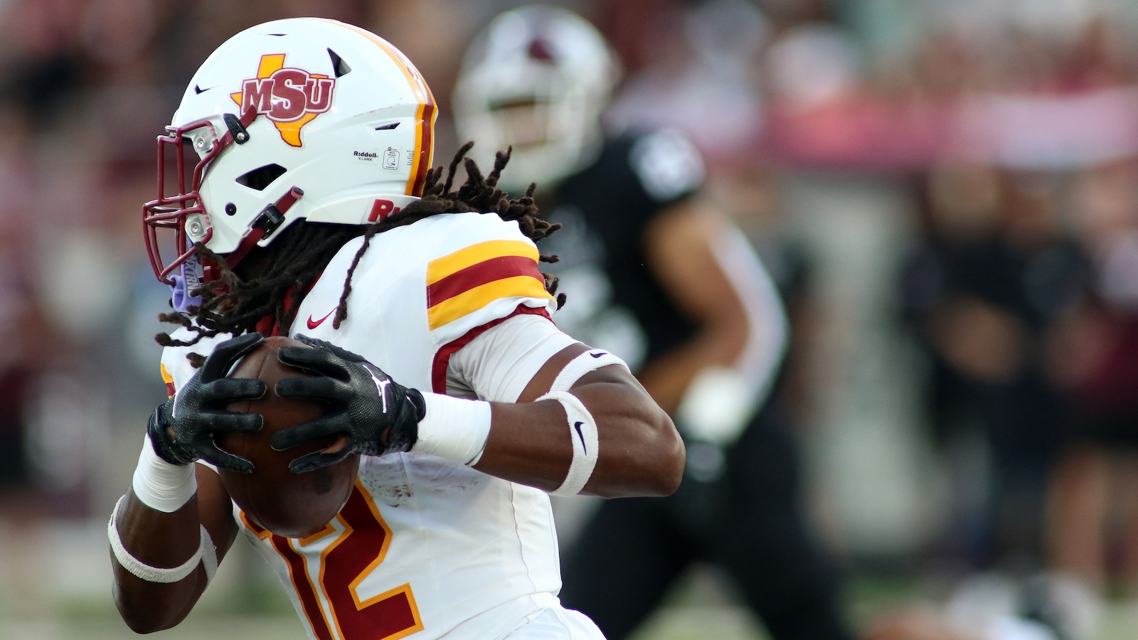 Mississippi State football player in a white jersey with red and gold stripes cradling the football during a game, helmet showing MSU logo.
