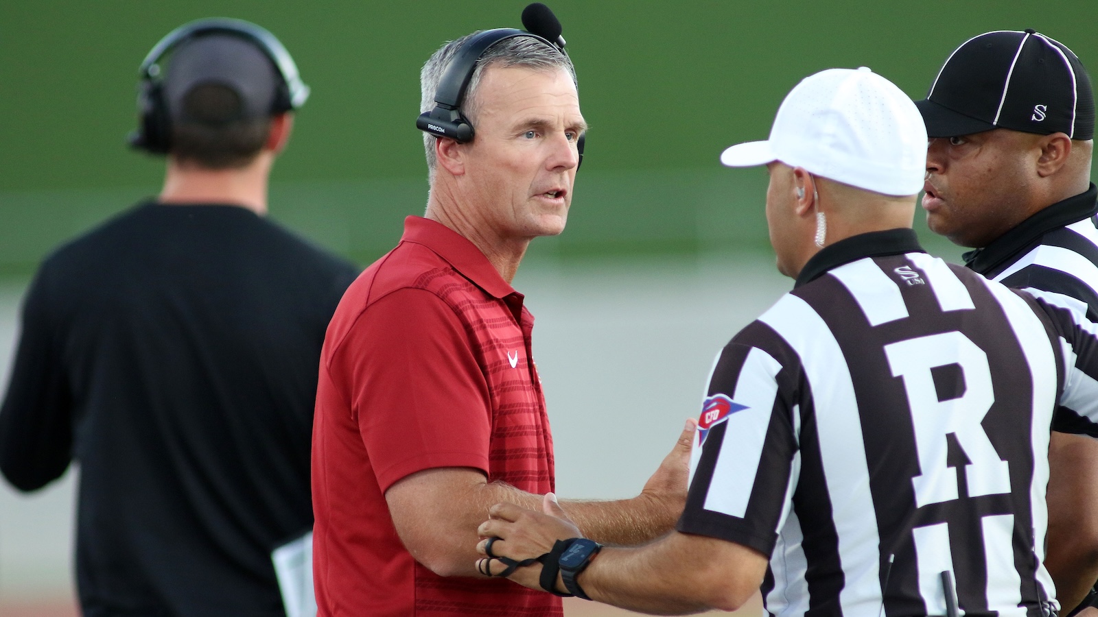 Football coach in a red polo talks with two referees in black-and-white stripes on the sideline, all wearing headsets.