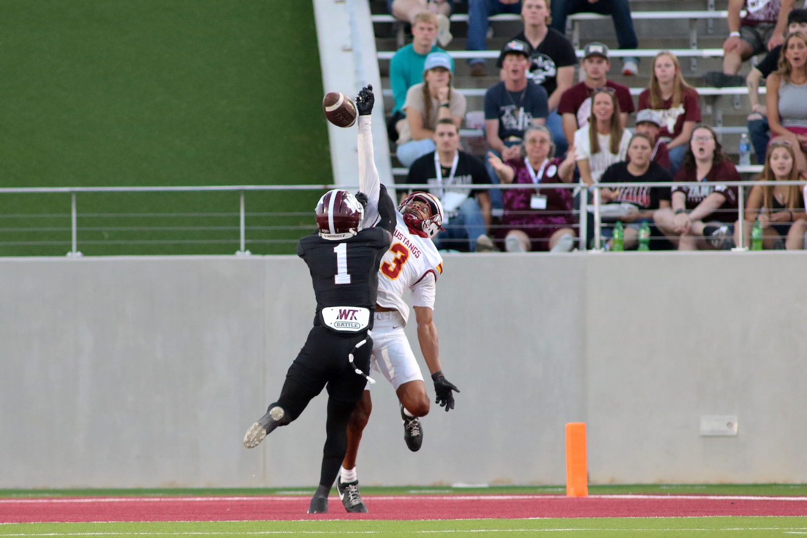 Football players jump for a pass near the end zone; a black jersey with number 1 blocks a white Mustangs player wearing number 3 while reaching for the ball as spectators watch from the stands behind the sideline.