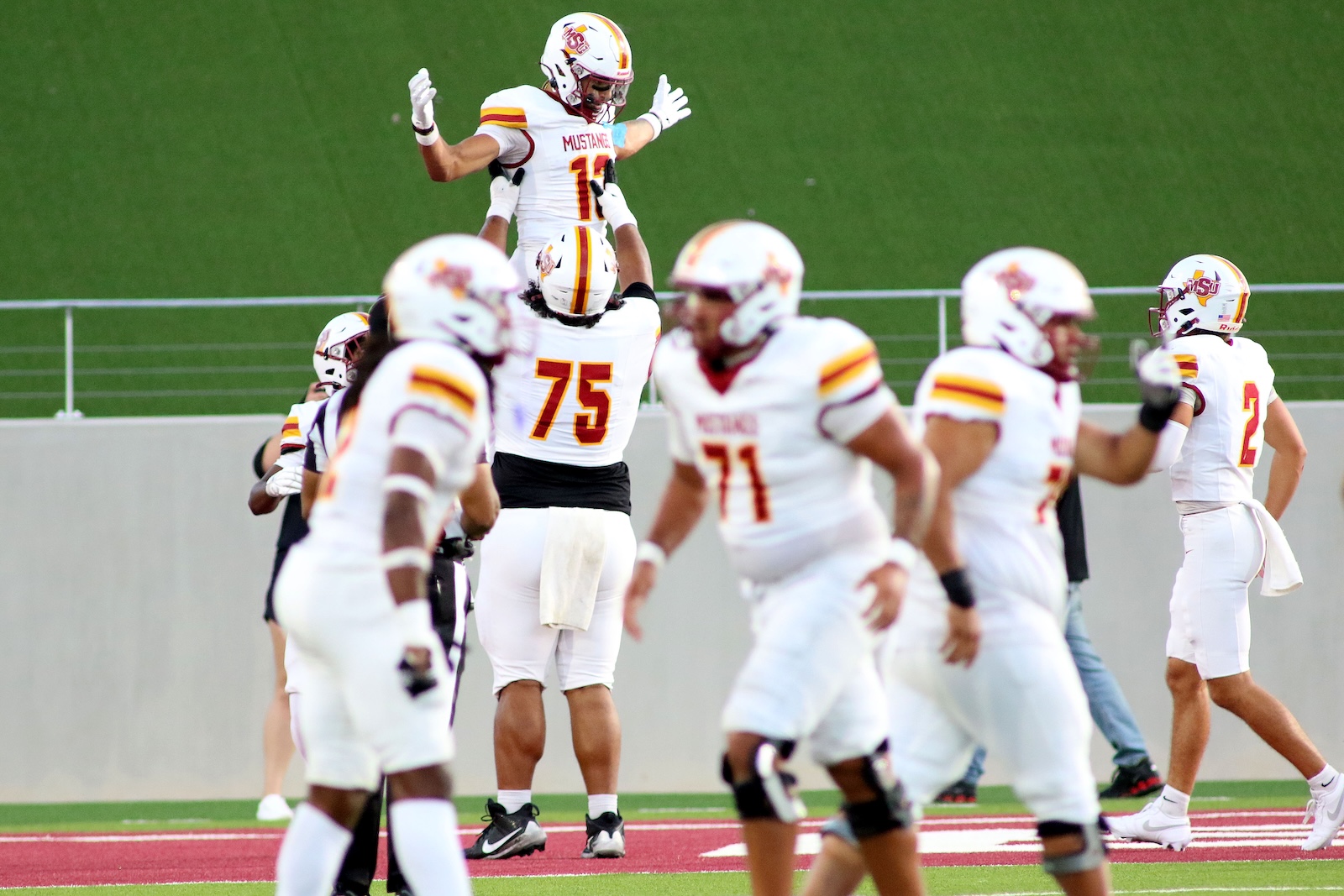 Football players in white uniforms celebrate a touchdown on the green turf as a teammate is lifted in the air by teammates in the foreground.