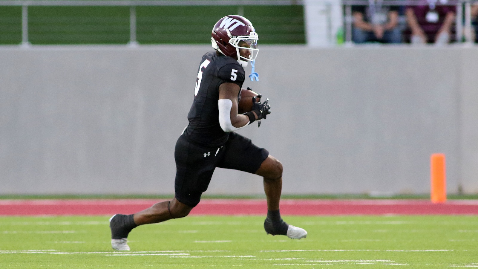 Football player in black uniform sprinting with ball toward the end zone on a green field.