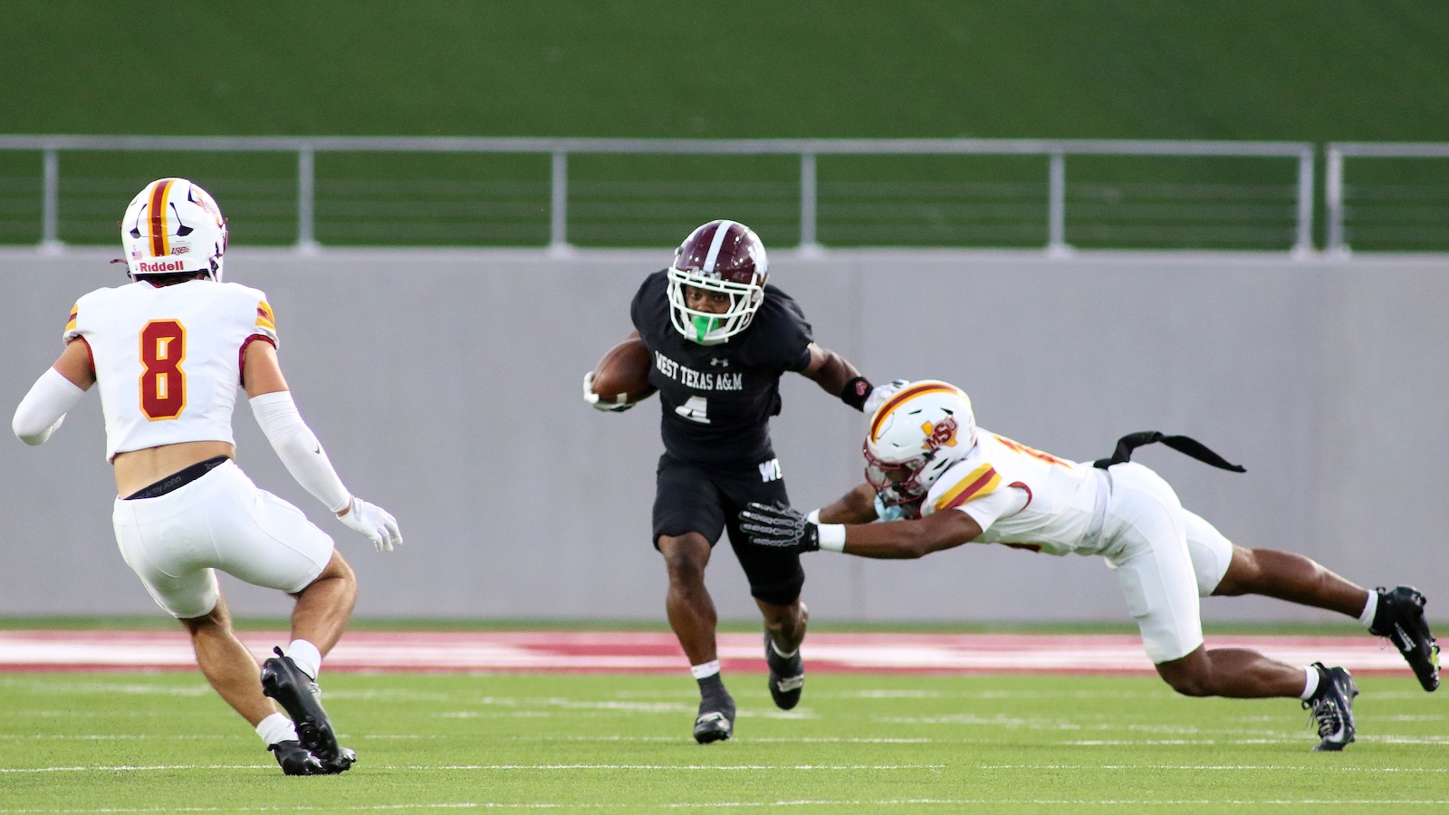 Football action: a ball carrier in a dark jersey runs with the ball while a defender in white dives to tackle him on the grass field.