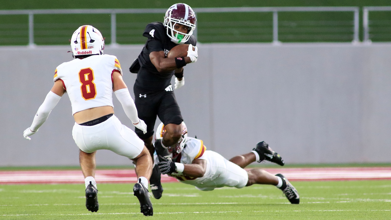 Football player in black uniform carries the ball as a defender in white (number 8) closes in on a tackle on a green field stadium backdrop.