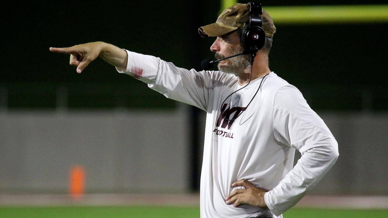 Football coach wearing a headset and camo cap points toward the field during a nighttime practice session on a football field.