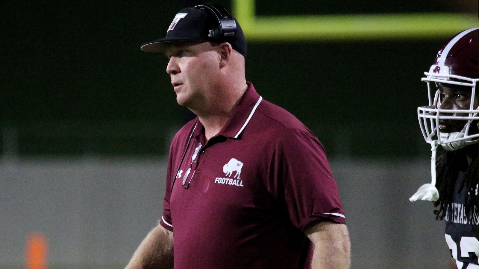 Football coach in a maroon polo with headset walking on the sideline, a player in a helmet beside him at dusk.