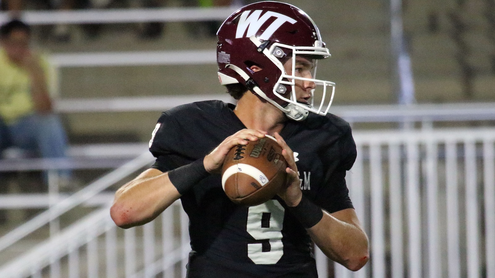 Quarterback in black jersey holding a football, ready to throw, wearing a maroon helmet with a white logo.