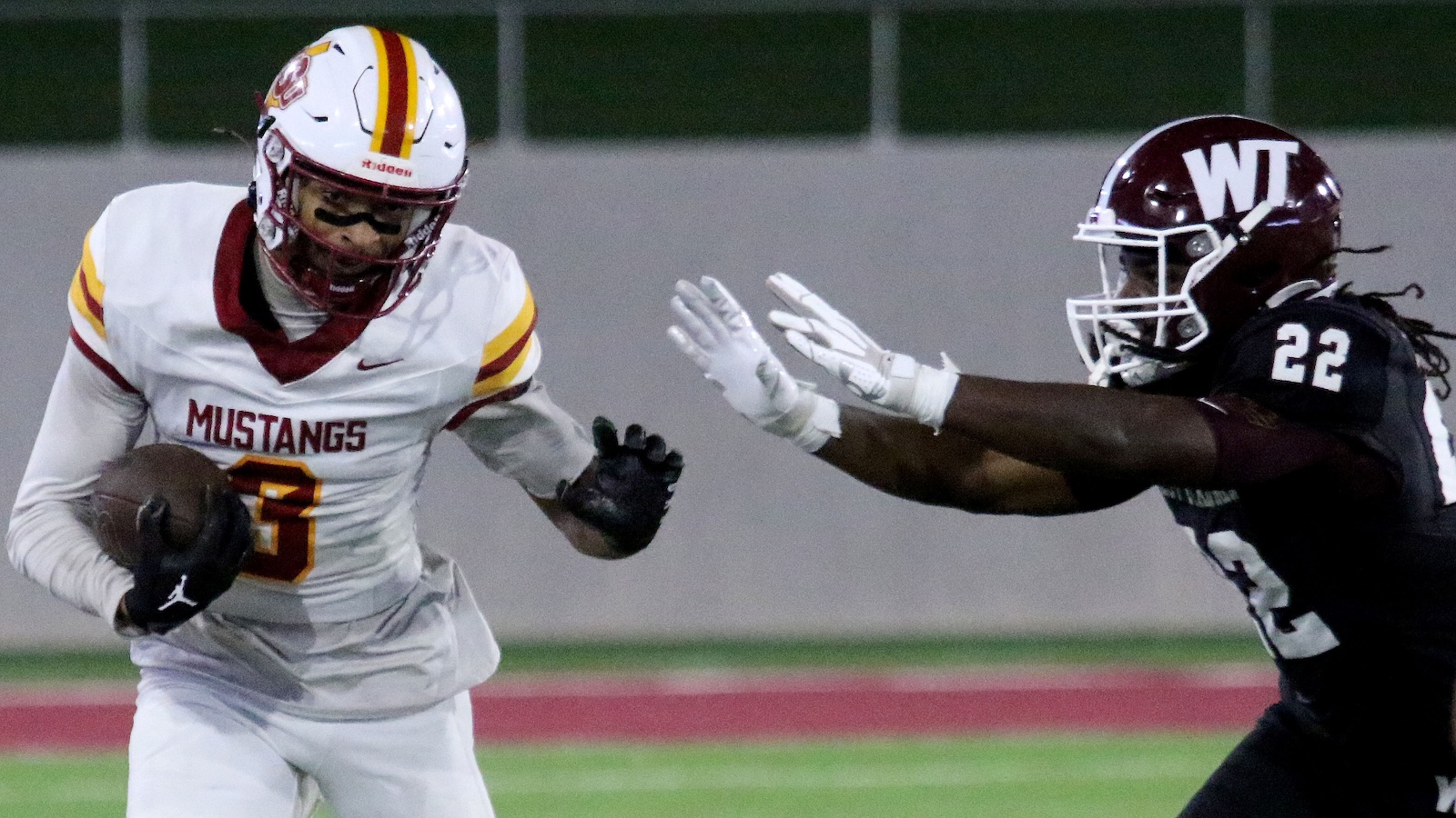 Football players in a high school game: a Mustangs ball carrier in white (number 3) runs with the ball, pursued by a WT defender (number 22) reaching to tackle.