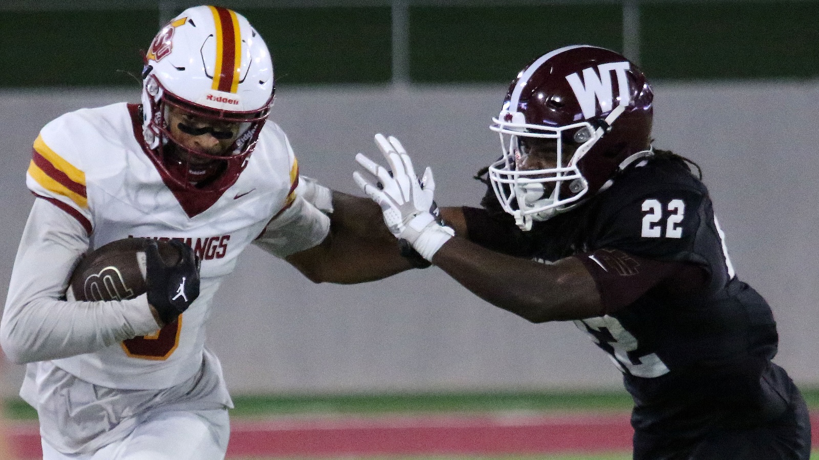 Football player in a white jersey runs with the ball while a defender in a dark uniform reaches in for a tackle.