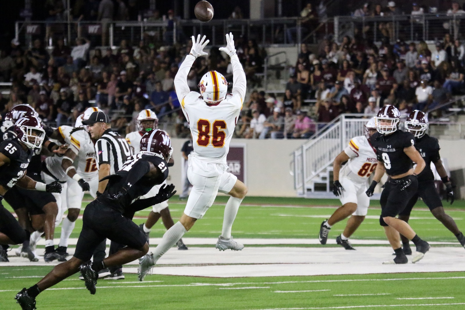 Football game: white-uniformed player (#86) leaps with arms raised to catch a pass as a ball descends, surrounded by black-uniformed players and a crowd.","Wide shot of a football game at night: #86 in white jumps to catch the ball while defenders in black pursue.","Athlete in white jersey #86 high-jumps to receive a pass during a crowded play on a lit field." ,"Action shot: linemen and a leaping receiver in white vie for a high pass as the ball hovers overhead." ,"In-game moment: player in white (#86) soars to reach the football amid players and spectators." ]} }```170```? Oops. Let's present proper JSON array. Remove stray. Final. → ````{