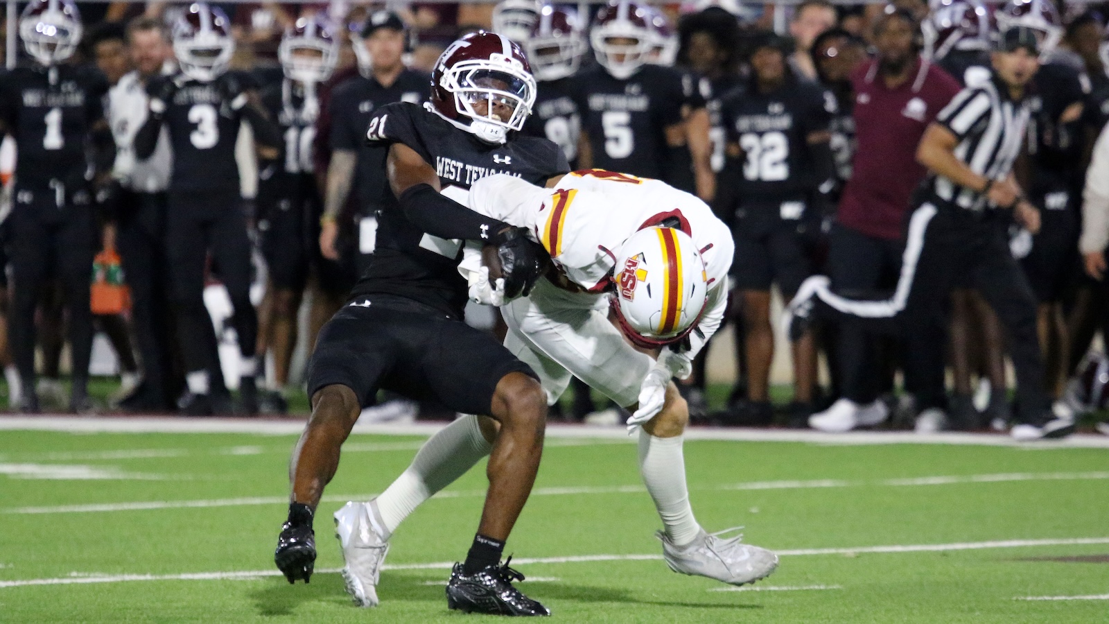 A football defender in a black West Texas uniform tackles a white-uniformed player during a game on a green turf field, with spectators on the sideline.