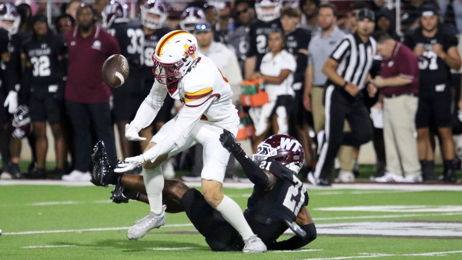 Football action: a player in white uniform leaps over a tackler in black as the ball sails through the air on a green field near the sideline crowd behind.