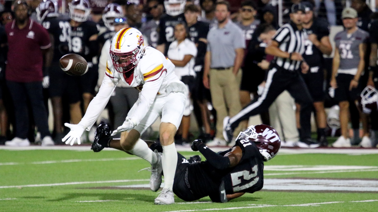 White-uniform football player extends for the ball as a tackle unfolds, ball in midair near him on a bright field with defenders in the background