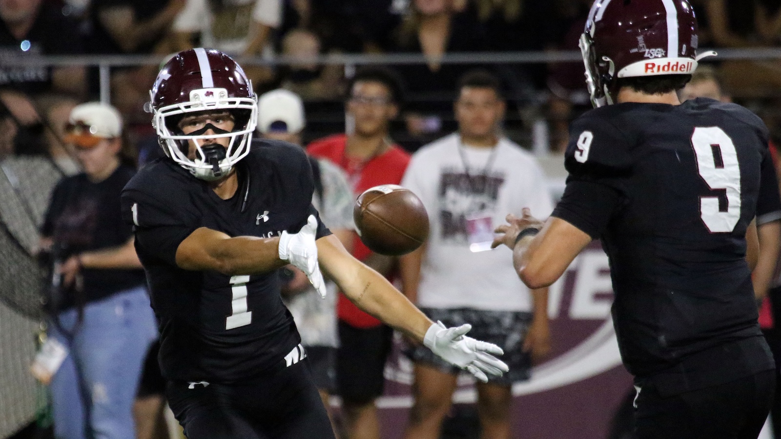 Two football players in black jerseys reach for a loose ball mid‑air, #1 extending a gloved hand while #9 moves in from the right.