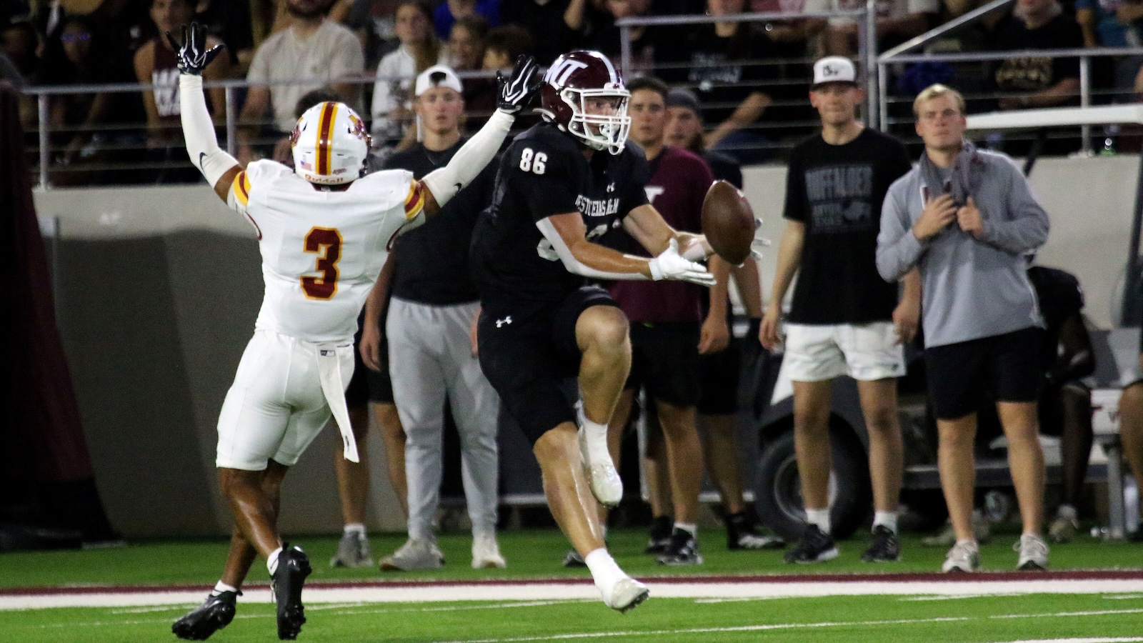Football players in a stadium: black jersey player (86) leaps to catch a pass while white-jersey defender (number 3) raises arms nearby. Crowded sideline in background.