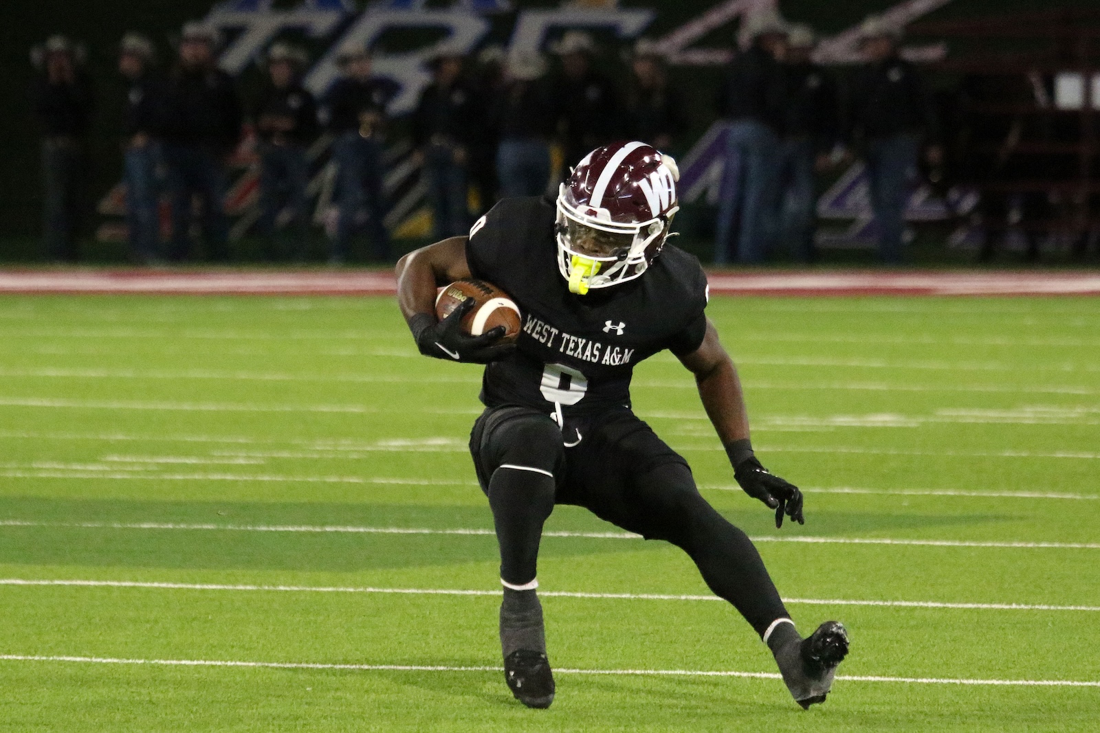 Football player in a black West Texas A&M uniform runs with the ball on a green turf field at night, helmet white with maroon stripe, yellow mouthguard visible.