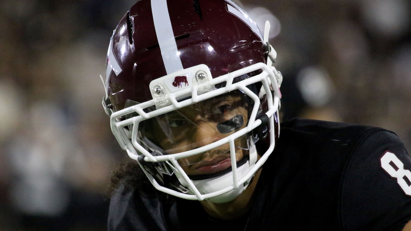 Close-up of a football player wearing a burgundy helmet with a white facemask, looking to the left with eye black under the eye.