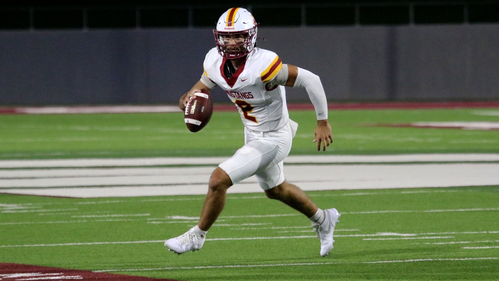 Football player in white uniform with a red and gold helmet rushing with the ball on a green turf field at night.