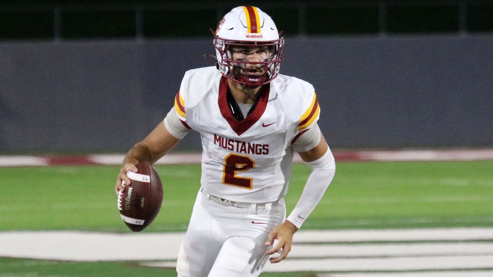 American football player in a white Mustang jersey with number 2, running with the ball on a green field at night stadium.