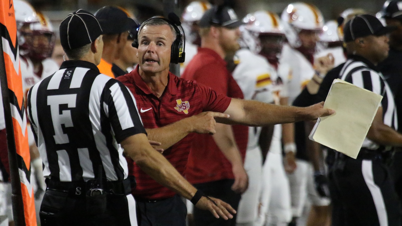 Football coach with headset points toward a clipboard held by an official on the sideline during a football game.