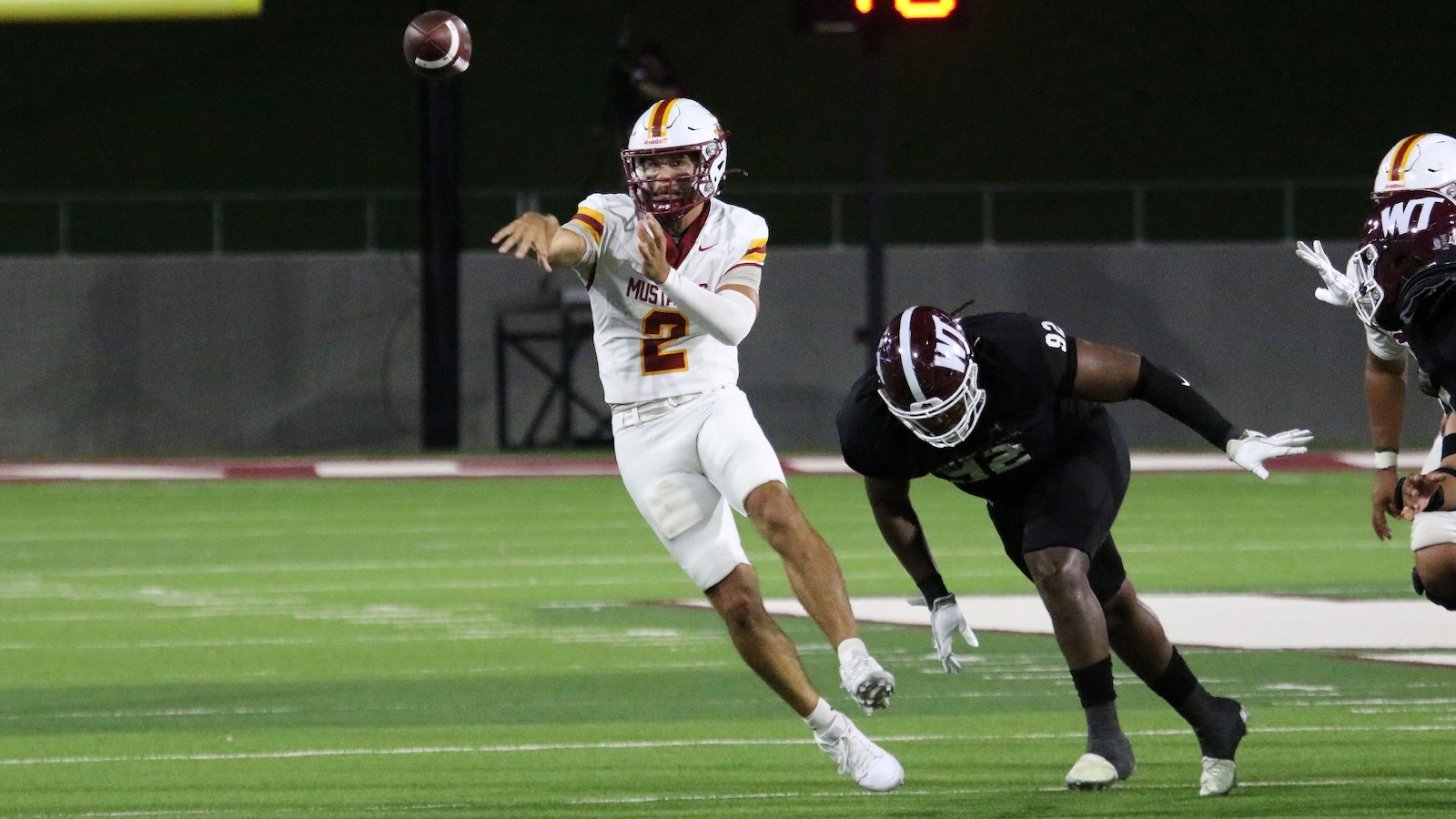 Quarterback in a white jersey with number 2 throws a pass as a defender in black lunges toward him during a night game.