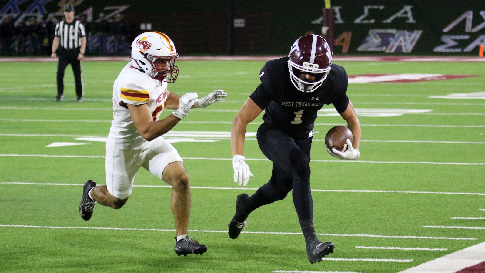 Football player in black West Texas A&M uniform runs with ball as defender in white MSU jersey reaches in.