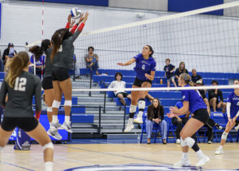 Blue team player #6 spikes the ball while two gray-uniformed players jump to block at the net during a volleyball match, with spectators watching from blue bleachers.