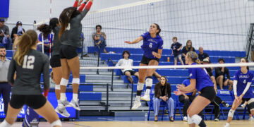 Blue team player #6 spikes the ball while two gray-uniformed players jump to block at the net during a volleyball match, with spectators watching from blue bleachers.