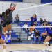 Blue team player #6 spikes the ball while two gray-uniformed players jump to block at the net during a volleyball match, with spectators watching from blue bleachers.