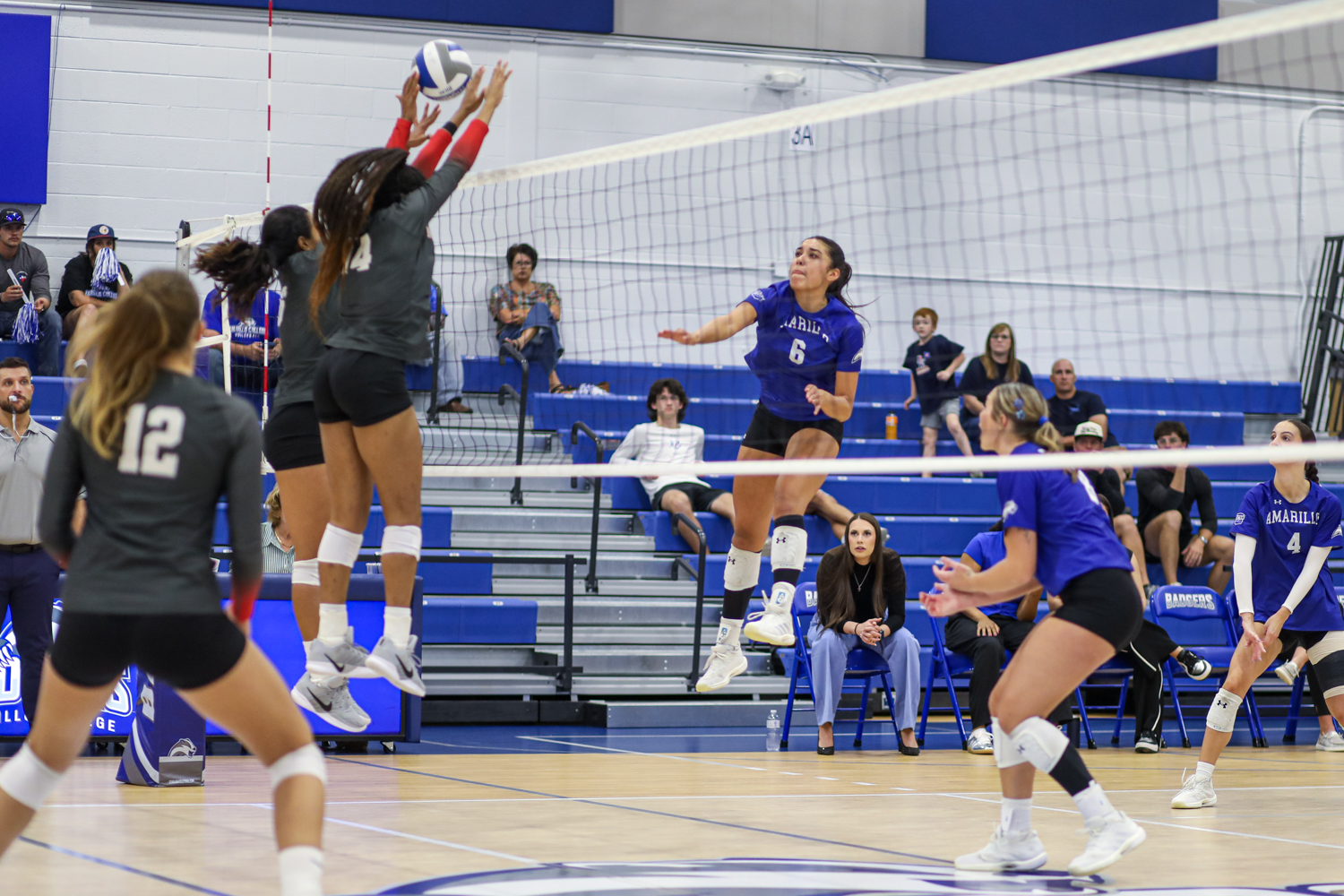 Edit 5 | Press Pass Sports Blue team player #6 spikes the ball while two gray-uniformed players jump to block at the net during a volleyball match, with spectators watching from blue bleachers.