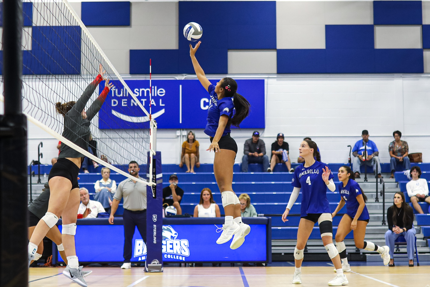 Edit 3 1 | Press Pass Sports Female volleyball player in a blue jersey jumps to spike the ball over the net while a teammate blocks at the net in a gym with blue bleachers behind her.