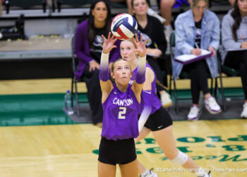 IMG_6125 | Press Pass Sports Two female volleyball players in purple uniforms reach up to set a ball during a match, with spectators watching in the background.