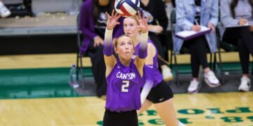 Two female volleyball players in purple uniforms reach up to set a ball during a match, with spectators watching in the background.