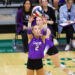 Two female volleyball players in purple uniforms reach up to set a ball during a match, with spectators watching in the background.