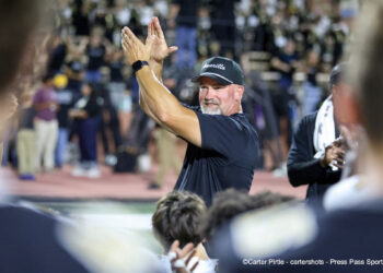 20260828-AmarilloHigh-PaloDuro-FB-045 | Press Pass Sports Man in a dark shirt and cap claps his hands in celebration on a sports field, with cheering spectators blurred in the background.