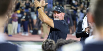 Man in a dark shirt and cap claps his hands in celebration on a sports field, with cheering spectators blurred in the background.