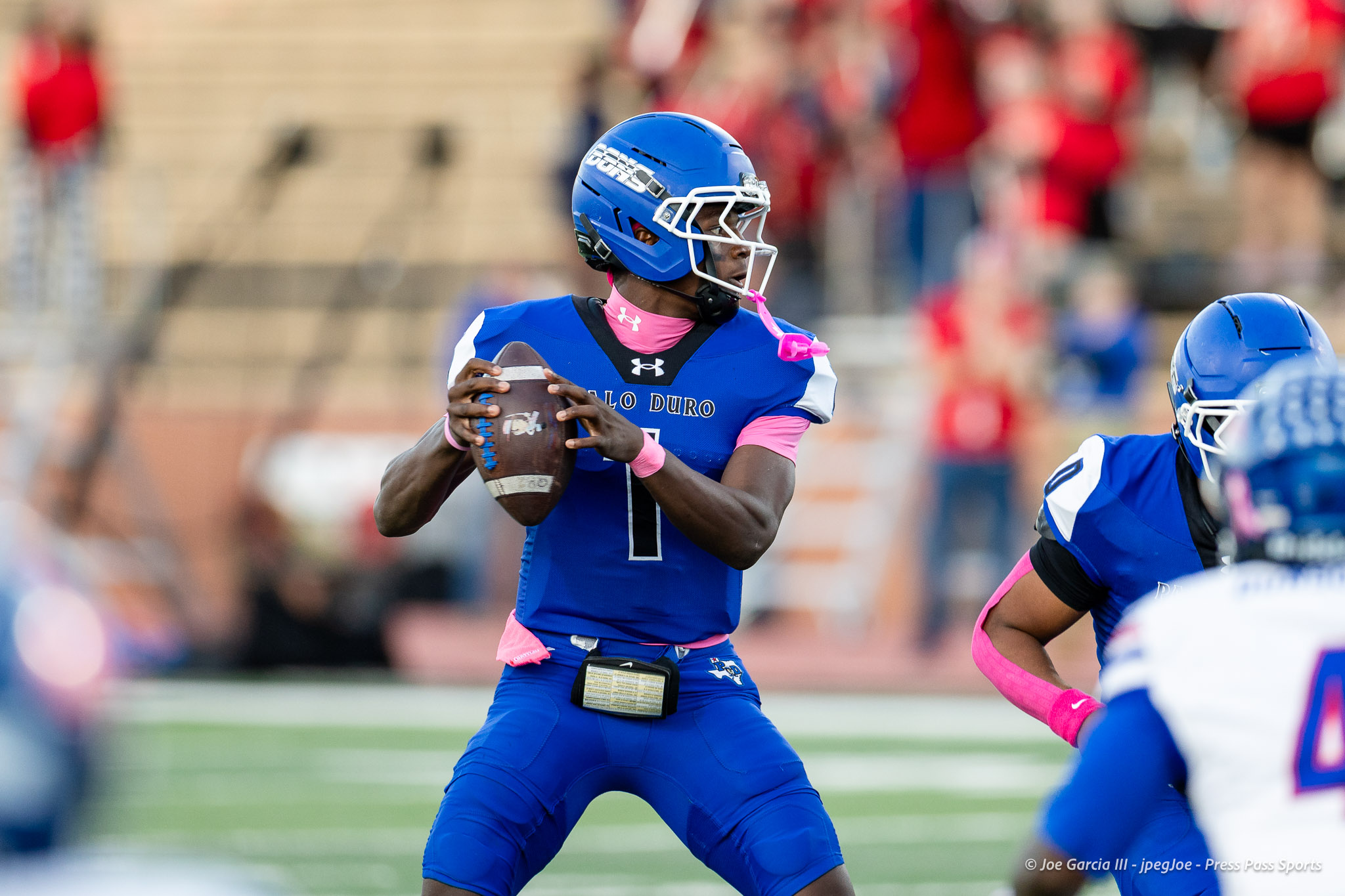 abilene-cooper-vs-palo-duro-jpegJoe-pps-2 | Press Pass Sports Quarterback in a blue and pink uniform prepares to throw a football on a crowded football field.