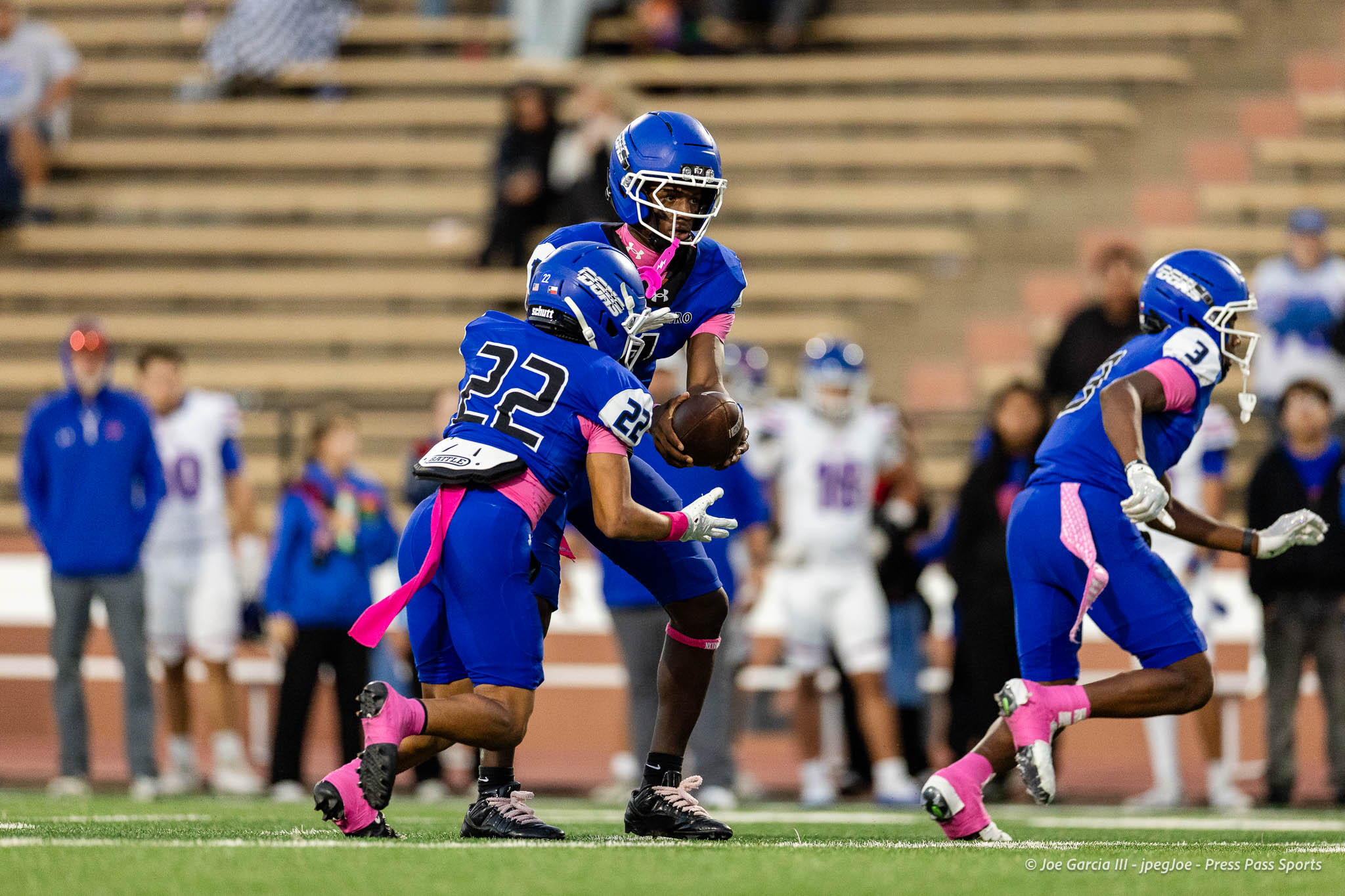 abilene-cooper-vs-palo-duro-jpegJoe-pps-13 | Press Pass Sports Blue-uniform football players run with the ball on a field during a game, with pink accents on their outfits and a stadium background.