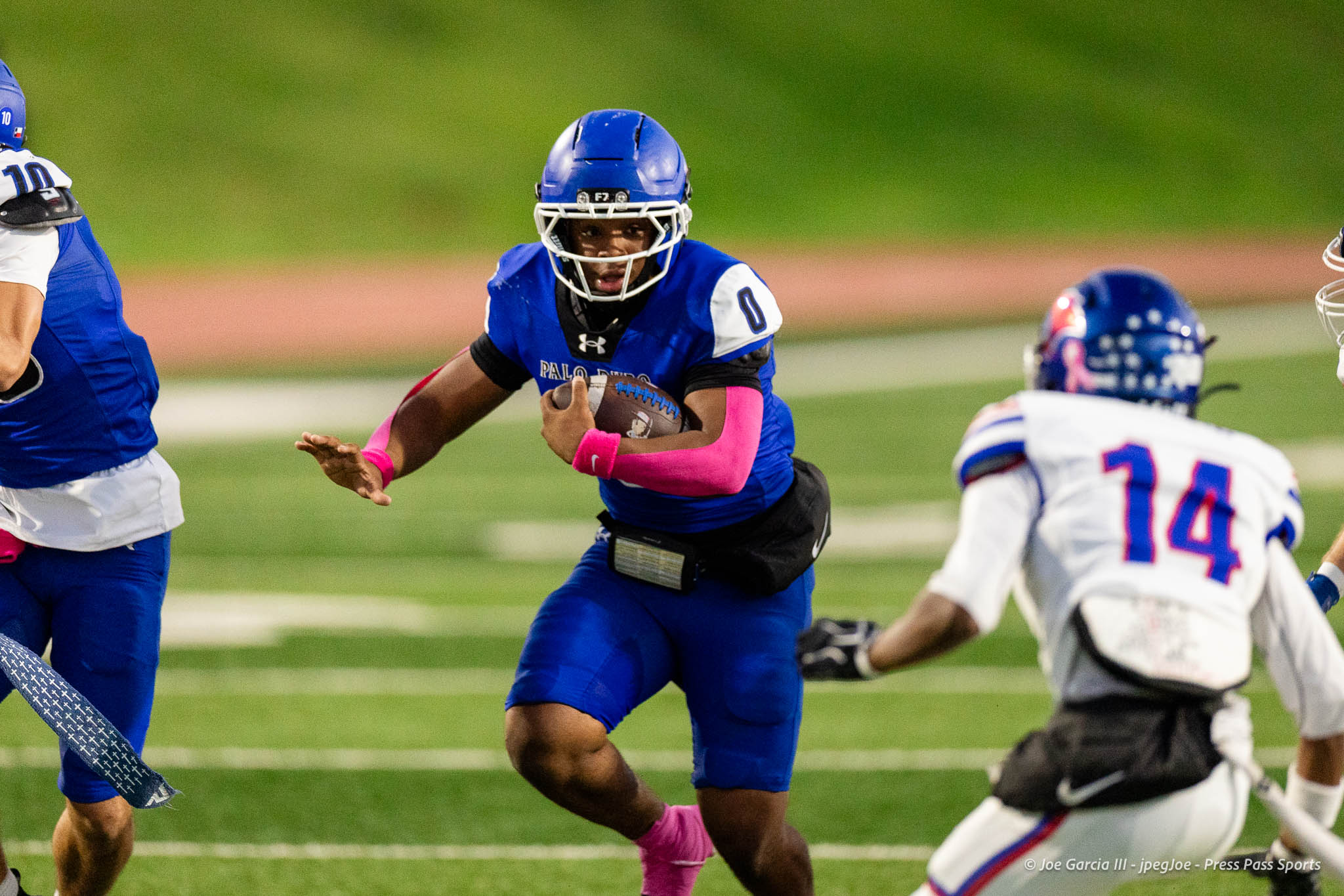 abilene-cooper-vs-palo-duro-jpegJoe-pps-14 | Press Pass Sports Football player in a blue uniform runs with the ball during a game on a green field, pink arm bands visible on the player.