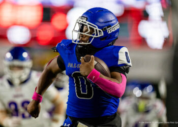 abilene-cooper-vs-palo-duro-jpegJoe-pps-18 | Press Pass Sports American football player in a blue uniform runs with the ball during a game, pink wristbands visible on his arms.