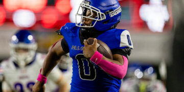 American football player in a blue uniform runs with the ball during a game, pink wristbands visible on his arms.
