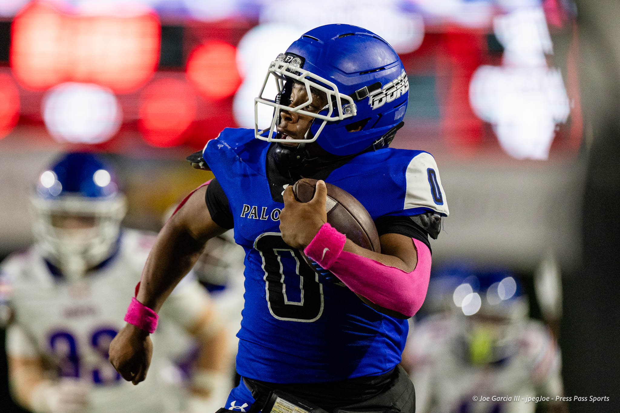 abilene-cooper-vs-palo-duro-jpegJoe-pps-18 | Press Pass Sports American football player in a blue uniform runs with the ball during a game, pink wristbands visible on his arms.