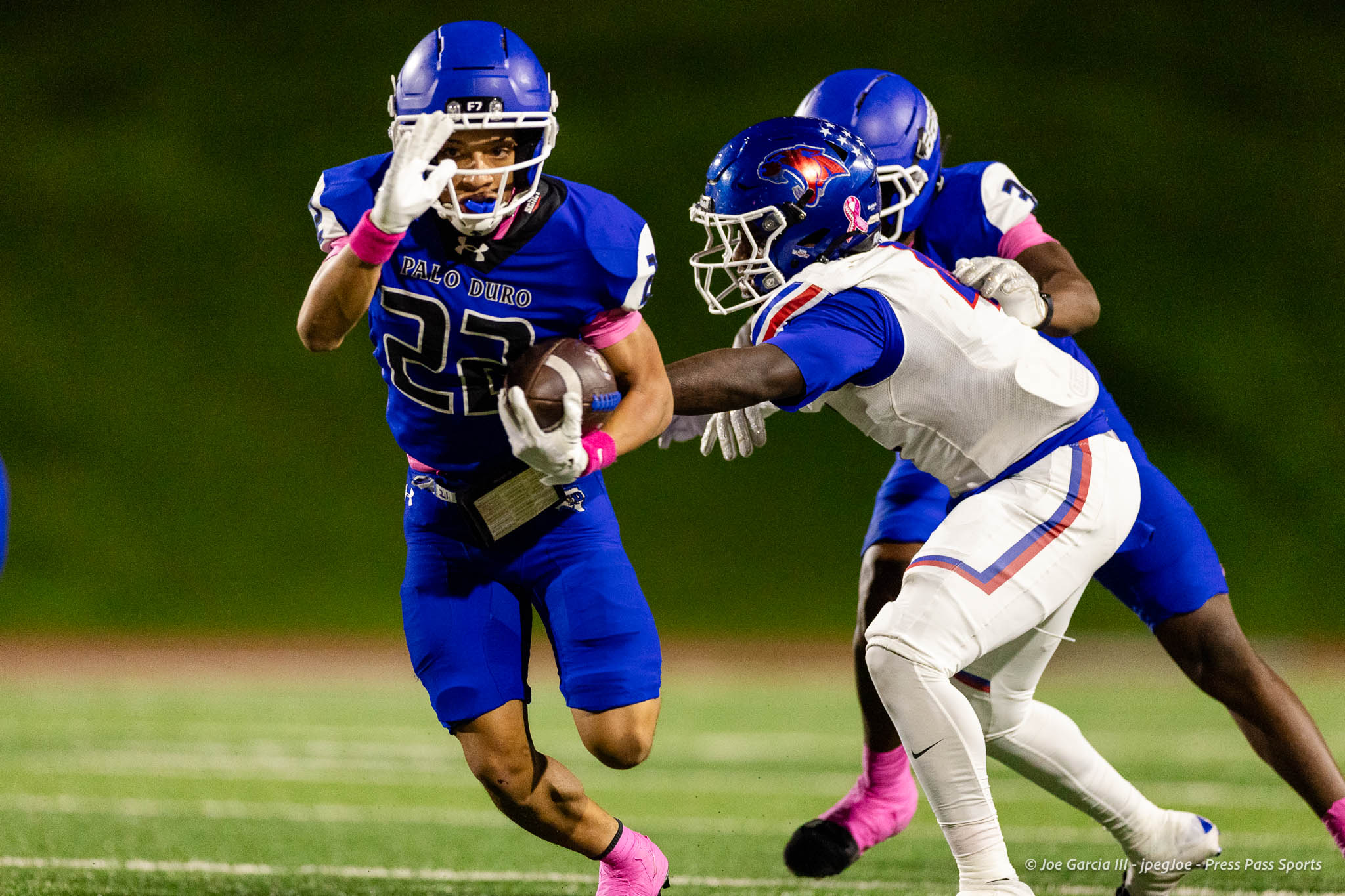 abilene-cooper-vs-palo-duro-jpegJoe-pps-21 | Press Pass Sports Palo Duro running back in blue jersey carries the ball while being chased by a defender in white, on a green turf under stadium lights, at night.
