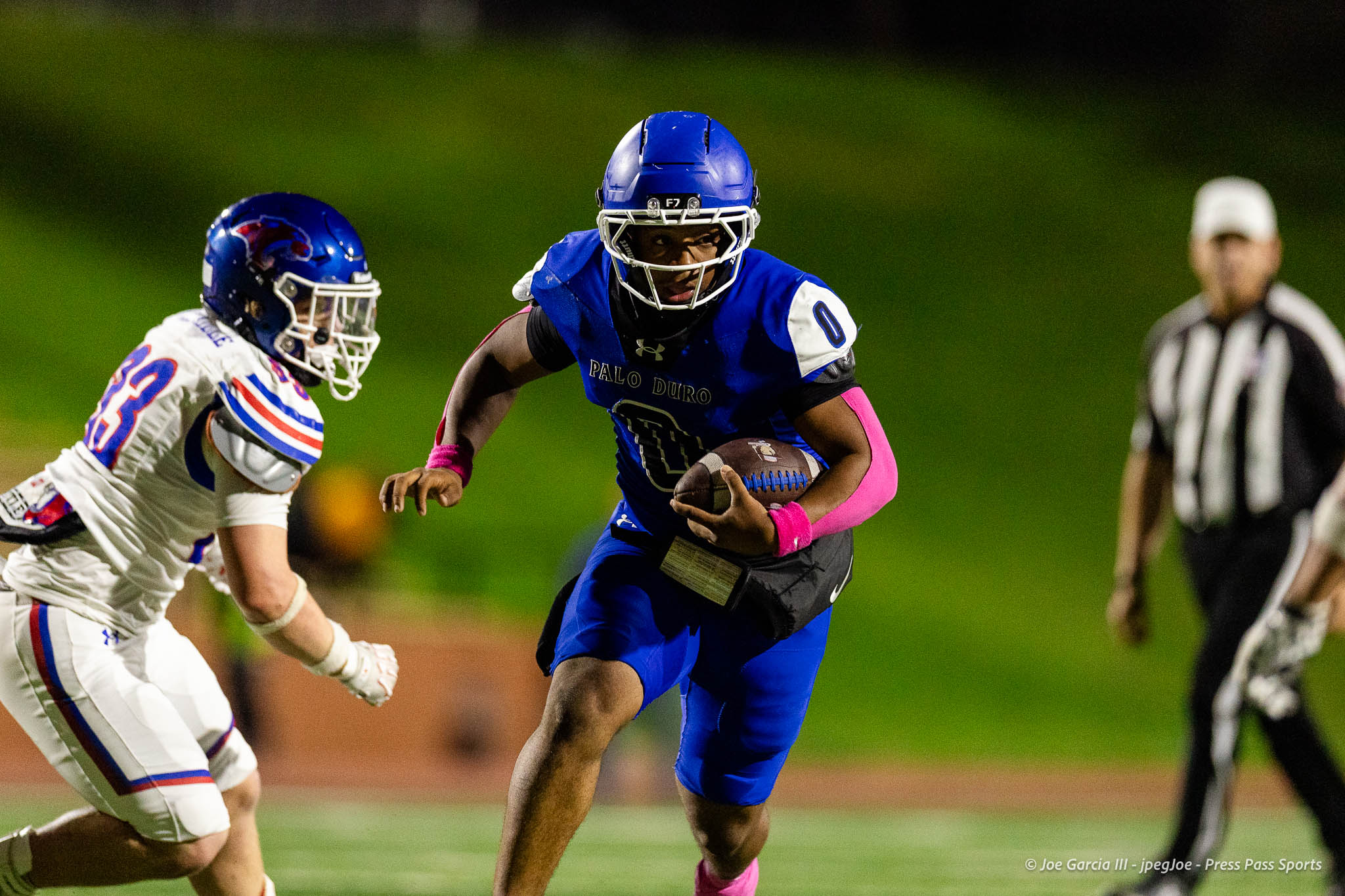 abilene-cooper-vs-palo-duro-jpegJoe-pps-22 | Press Pass Sports Blue-uniform football player with the ball sprinting downfield, pursued by a white-uniform defender on a green field, referee in background.