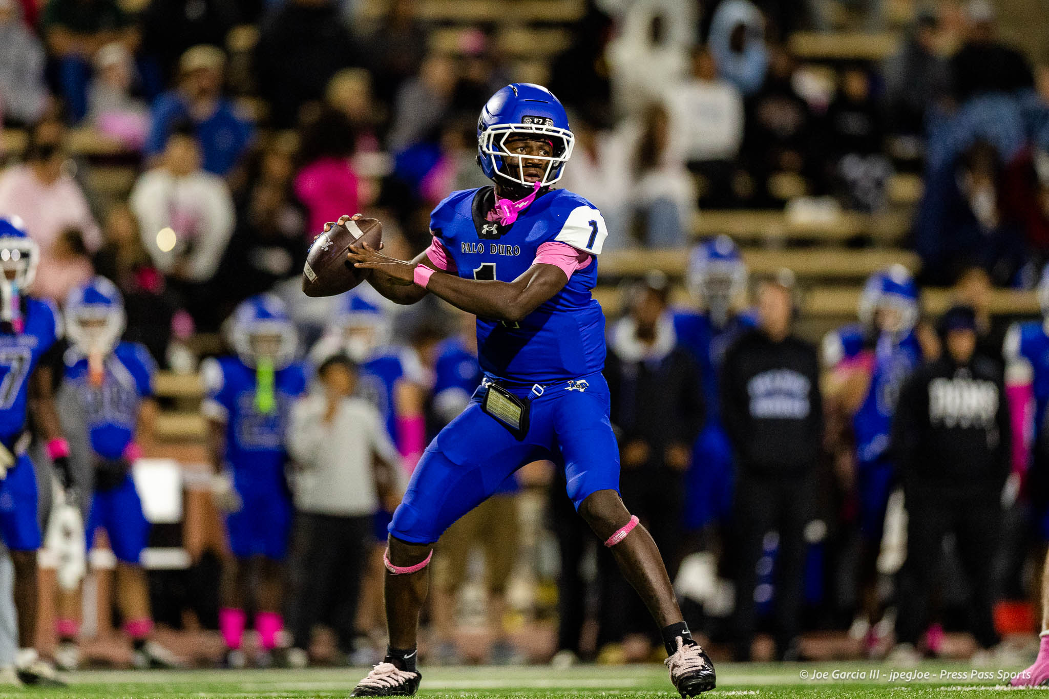abilene-cooper-vs-palo-duro-jpegJoe-pps-25 | Press Pass Sports Football quarterback in a blue uniform (#1) preparing to throw a pass during a game with a crowded stands backdrop.