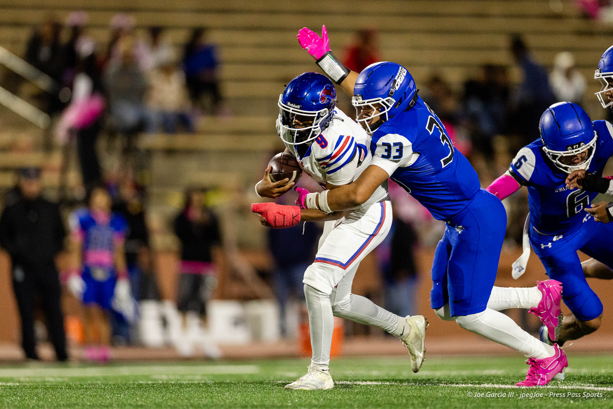 abilene-cooper-vs-palo-duro-jpegJoe-pps-26 | Press Pass Sports Football game action: a white-jersey player runs with the ball as blue-jersey defenders close in, pink gloves visible in a tackle attempt